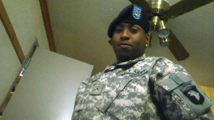 A man wearing Army fatigues and a beret with a blue crest on it. There is a patch on the shoulder. He is in a plain room with a ceiling fan above him and an open door behind him.