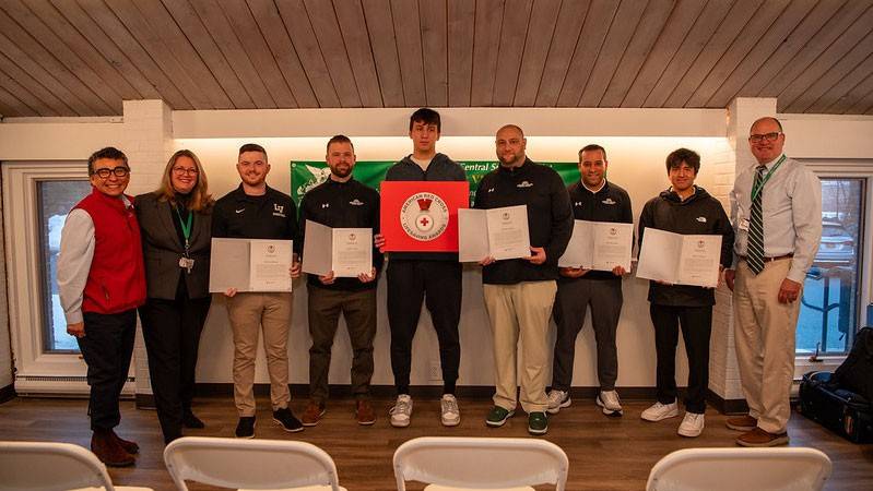 A group of adults stand indoors holding certificates, with one holding a certificate with a Red Cross insignia, in front of a green school district banner.
