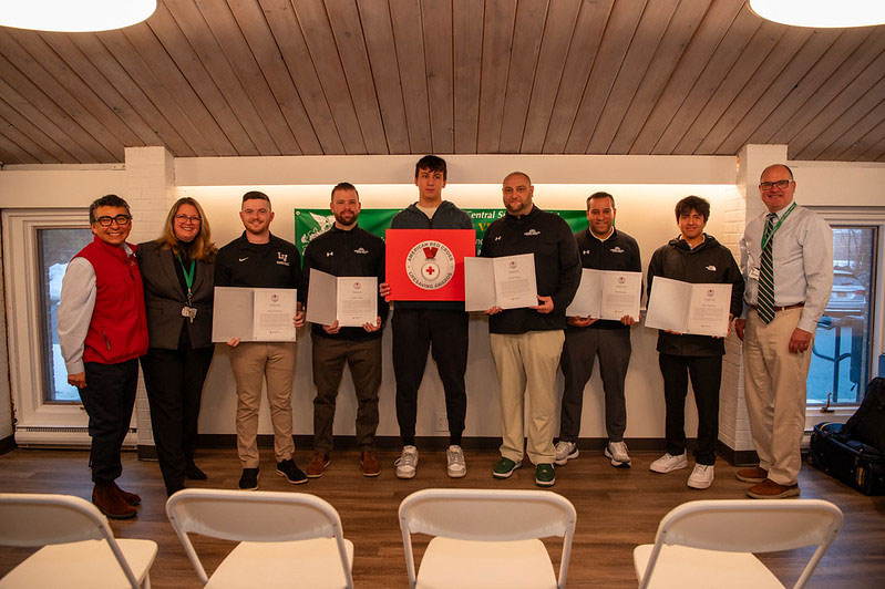 A group of adults stand indoors holding certificates, with one holding a certificate with a Red Cross insignia, in front of a green school district banner.