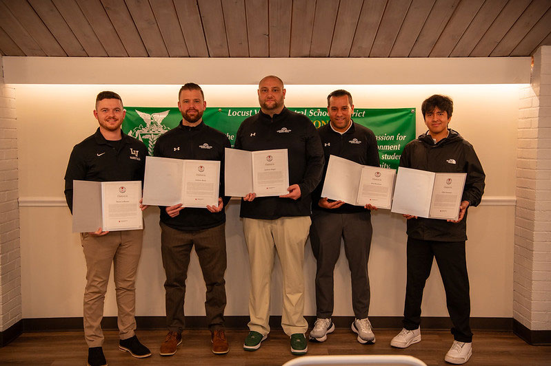 Five adults stand indoors holding certificates in front of a green Locust Valley Central School District banner.