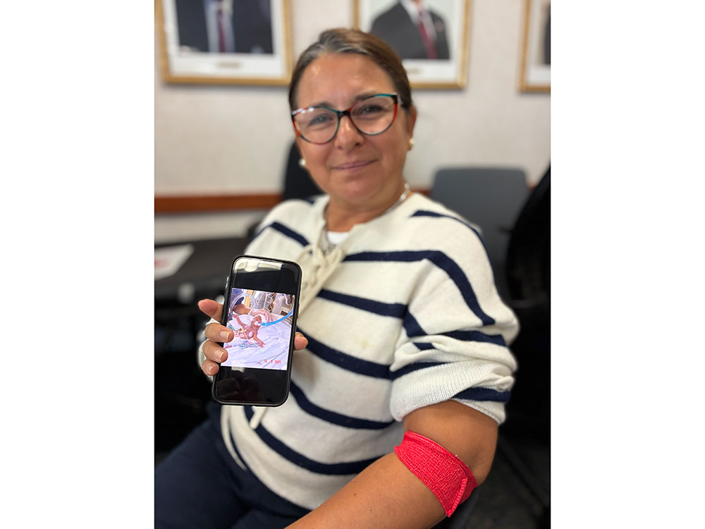 A seated person in a white and navy striped sweater, with a red blood donation arm band wrapped around her elbow, holds a cell phone showing a photo of a newborn baby connected to tubes and machinery.