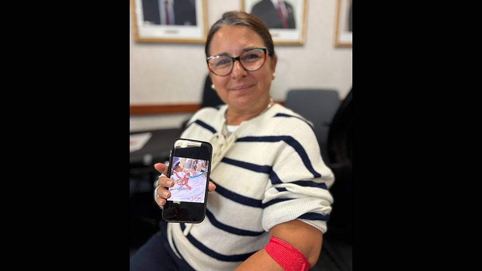 A seated person in a white and navy striped sweater, with a red blood donation arm band wrapped around her elbow, holds a cell phone showing a photo of a newborn baby connected to tubes and machinery.