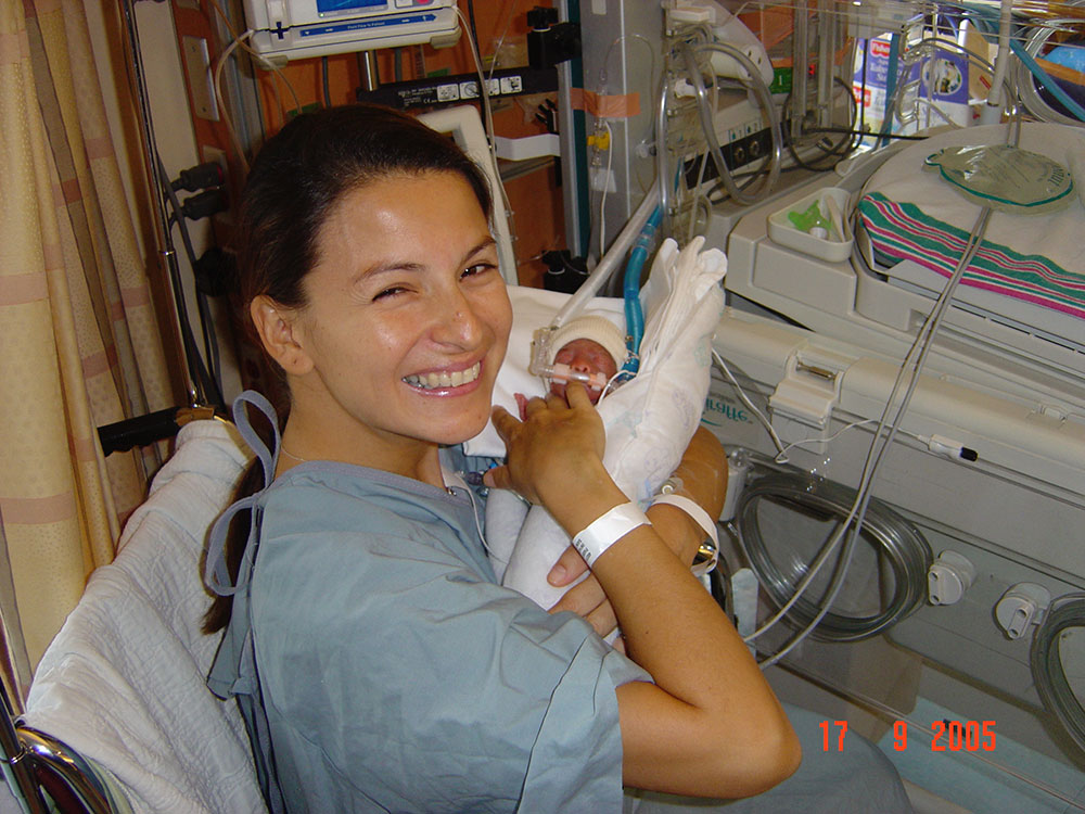 A smiling mother wearing a hospital gown holds a newborn baby that is connected to tubes and machines. There is an orange date in the lower corner which says 17 9 2005.