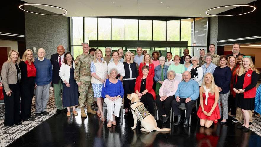 large group photo of Red Cross volunteers, military members and a service dog inside a building.
