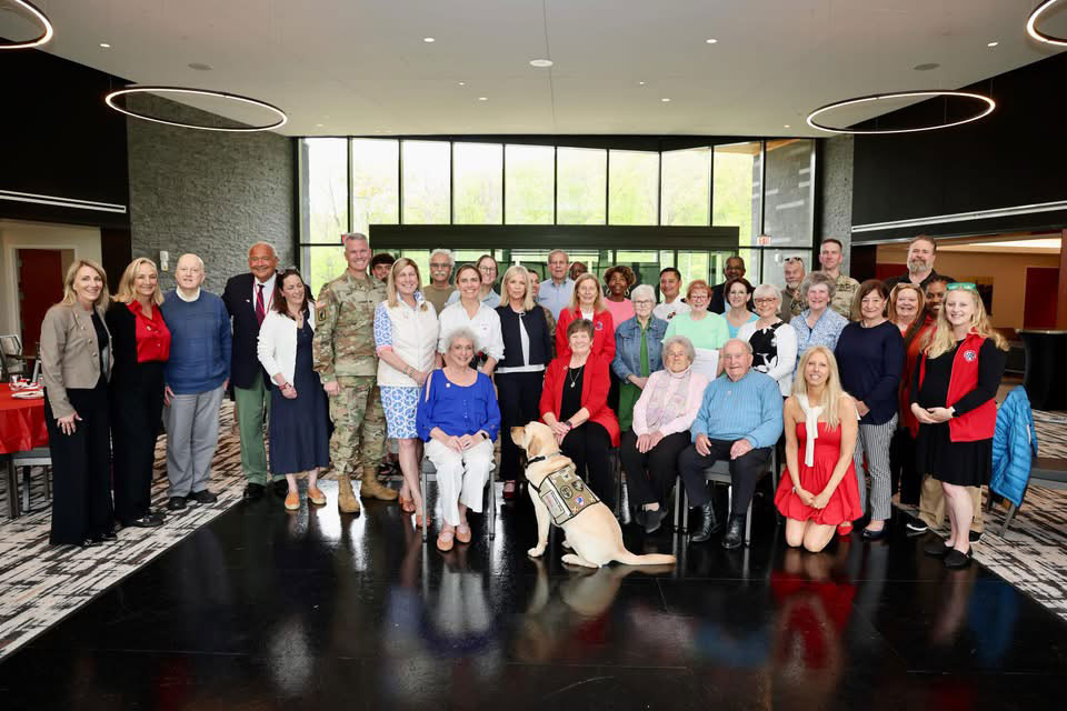 large group photo of Red Cross volunteers, military members and a service dog inside a building.