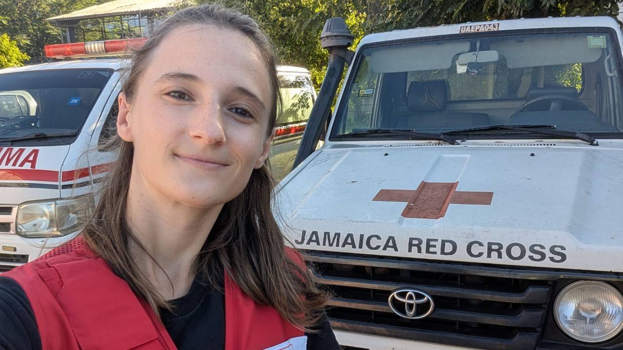 Person with long brown hair smiling and wearing an American Red Cross vest standing in front of two vehicles, including one marked Jamaica Red Cross.