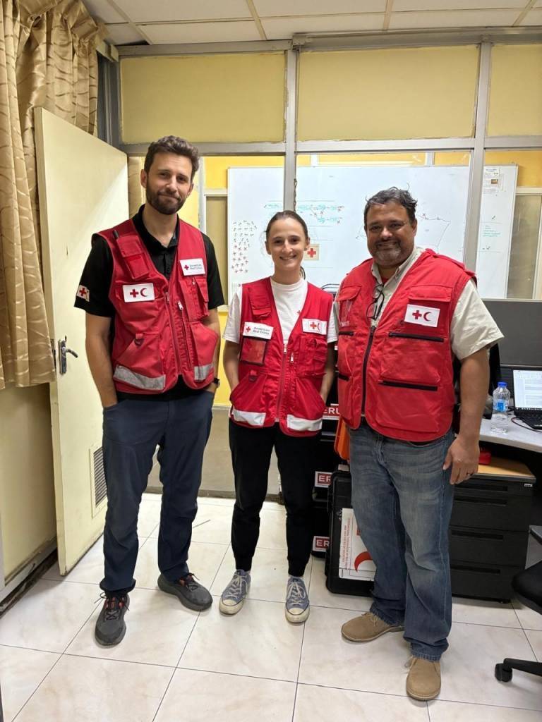 Three people wearing vests with logos that show both a red cross and a red crescent standing together in an office setting..