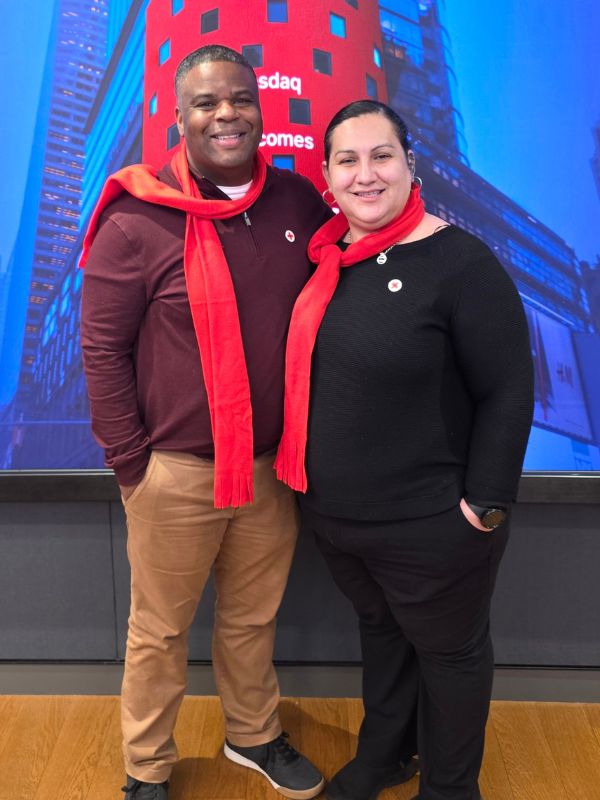Two people wearing red scarves and Red Cross pins stand smiling in front of a large NASDAQ display.