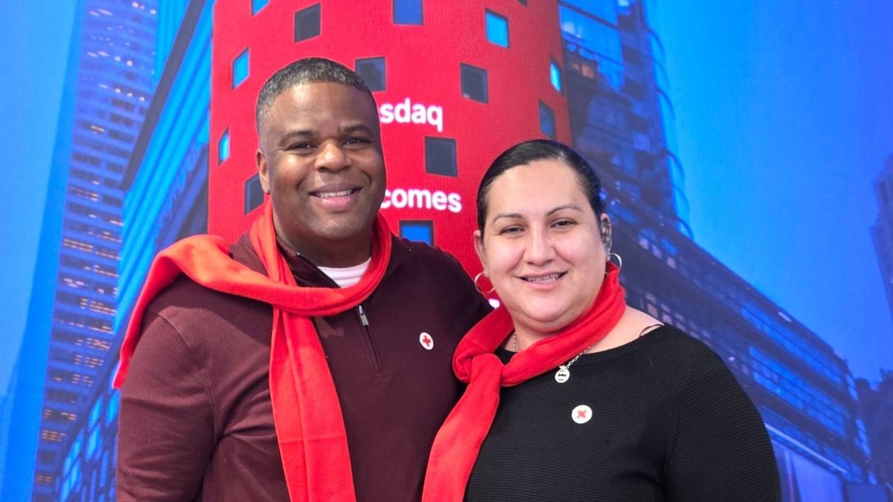 two people smiling in front of a large NASDAQ display