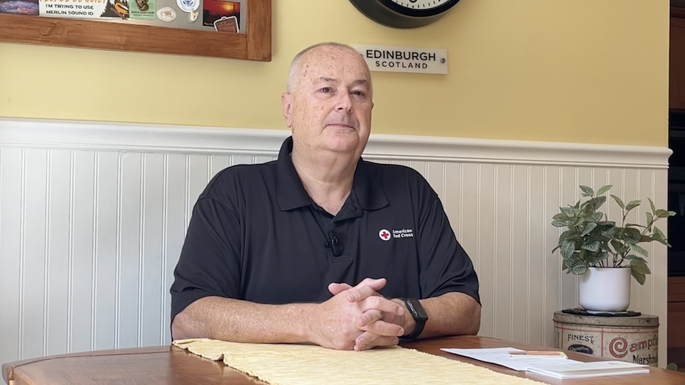Adrian Grieve wearing a black polo shirt with an American Red Cross logo, sitting at a table with his hands folded.