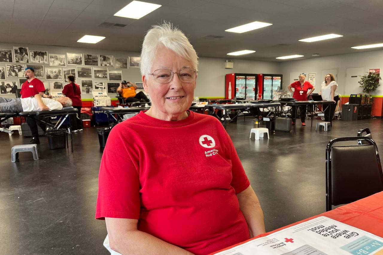 Carol Eroh sitting at a table wearing a red shirt with an American Red Cross logo. A blood drive is visible in the background. 
