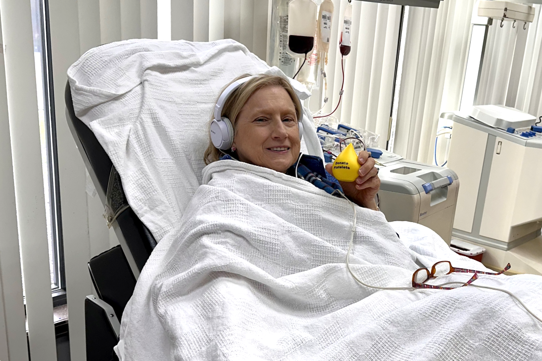Caroline Howell sits in a blood donation chair and holds a yellow blood drop top with the text "donate platelets". She is wearing headphones and is draped in a white blanket. Blood donation equipment is visible in the background.
