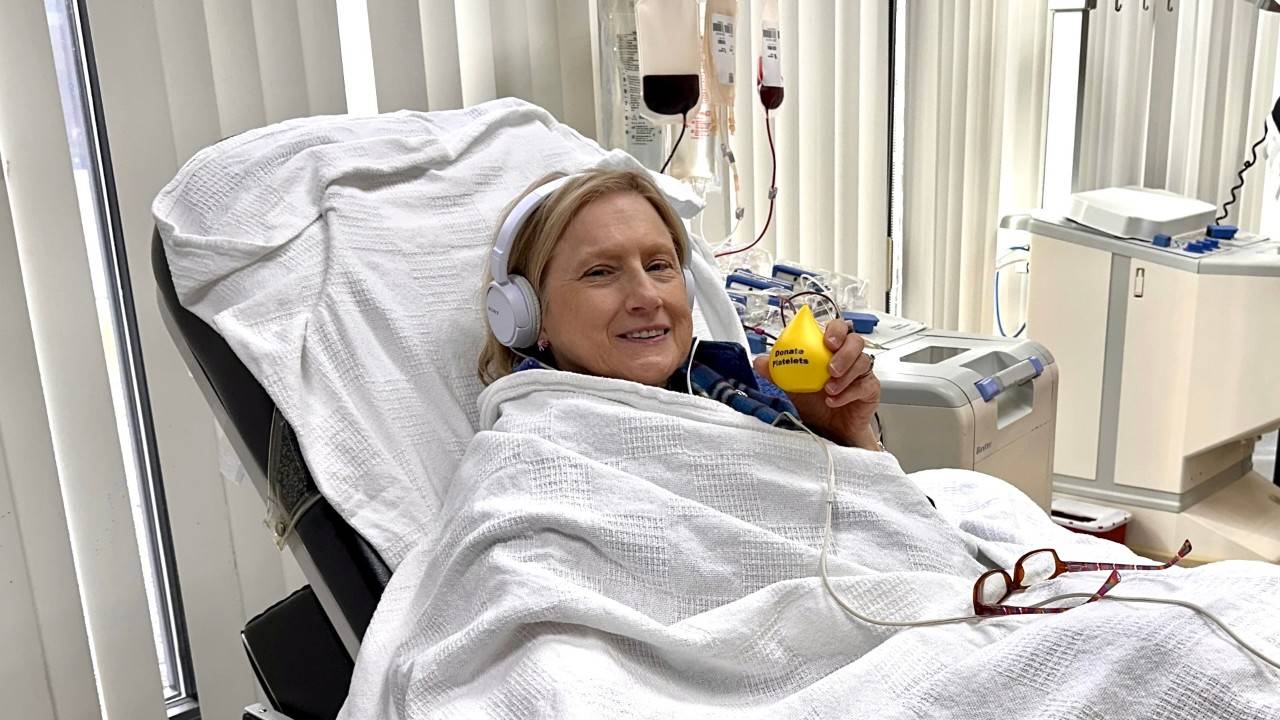 Caroline Howell in a platelet donation chair, holding a yellow blood drop toy that says "Donate Platelets". Caroline is wearing headphones and is draped in a white blanket. Platelet donation equipment is visible in the background.