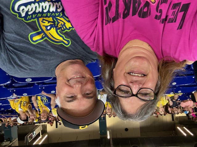 Catherine and David Truckley smiling at a baseball game. Catherine is wearing a pink shirt and David is wearing a gray shirt with a Savannah Bananas logo.