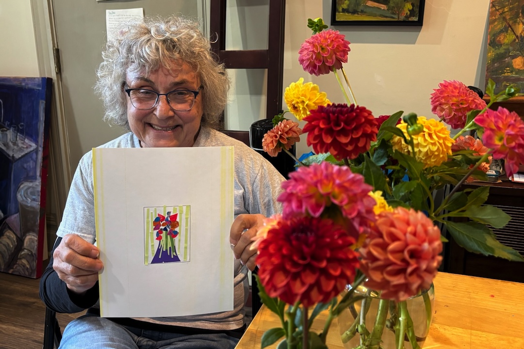Debra Kindervatter, smiling while holding a painting of flowers. A vase of flowers is on the table besides her.