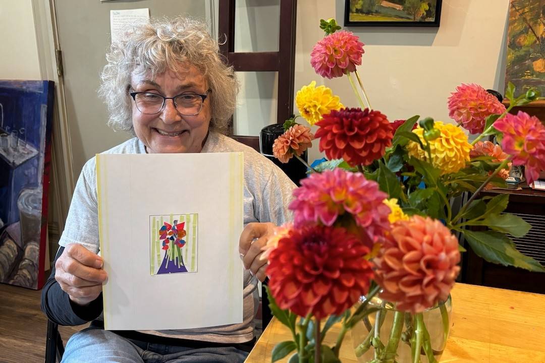 Debra Kindervatter, smiling while holding a painting of flowers. A vase of flowers is shown besides her.