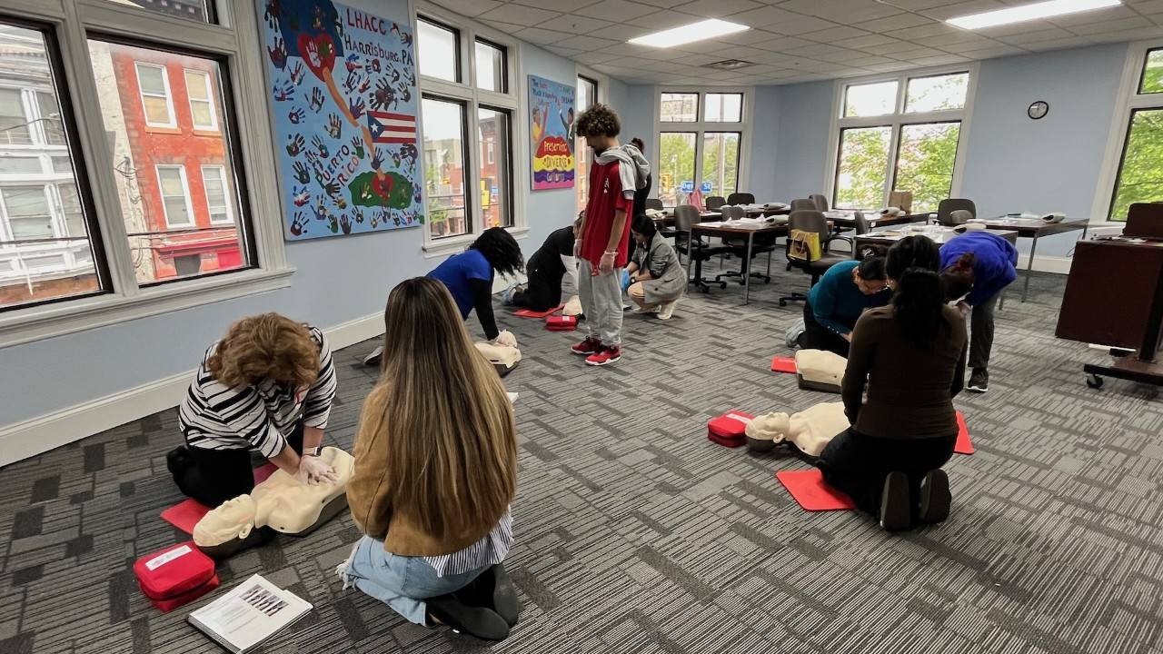 Class participants kneel on the floor next to CPR manikins. A mural on the wall says "LHACC Harrisburg" and shows the Puerto Rican flag.