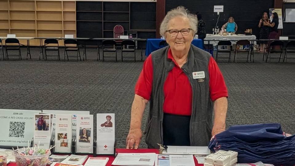 A person in a red shirt and a Red Cross name tag smiling behind a table filled with informational pamphlets.