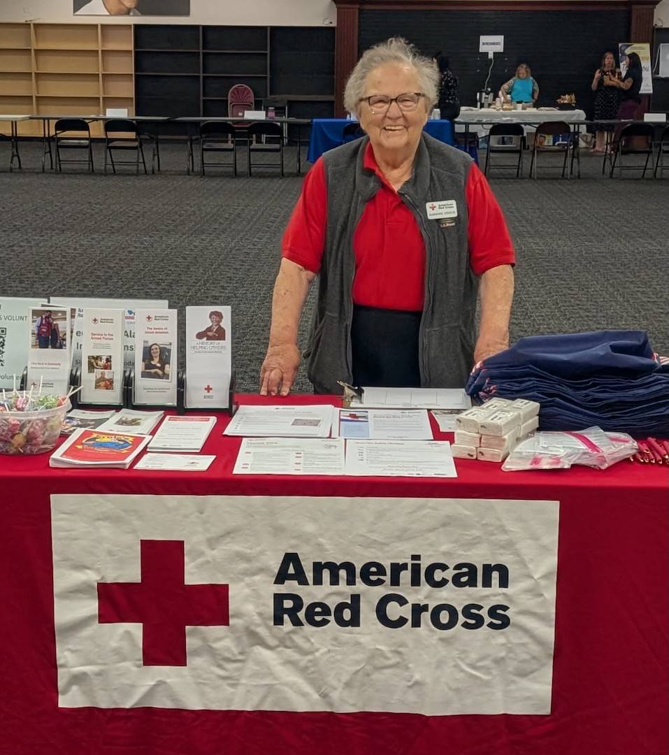 Marianne Konior wearing a red shirt and an American Red Cross name tag, standing behind a table filled with informational pamphlets. An American Red Cross tablecloth is on the table.