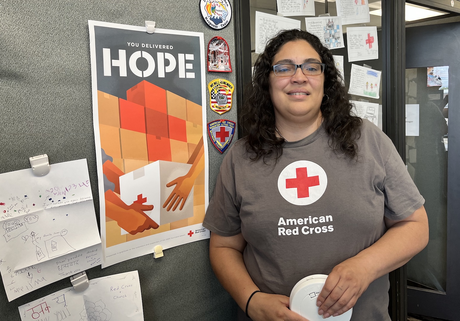 Rosalie Gonzalez in a gray Red Cross t-shirt, holding a smoke alarm, smiling in front a of a poster that says "You Deliver Hope"