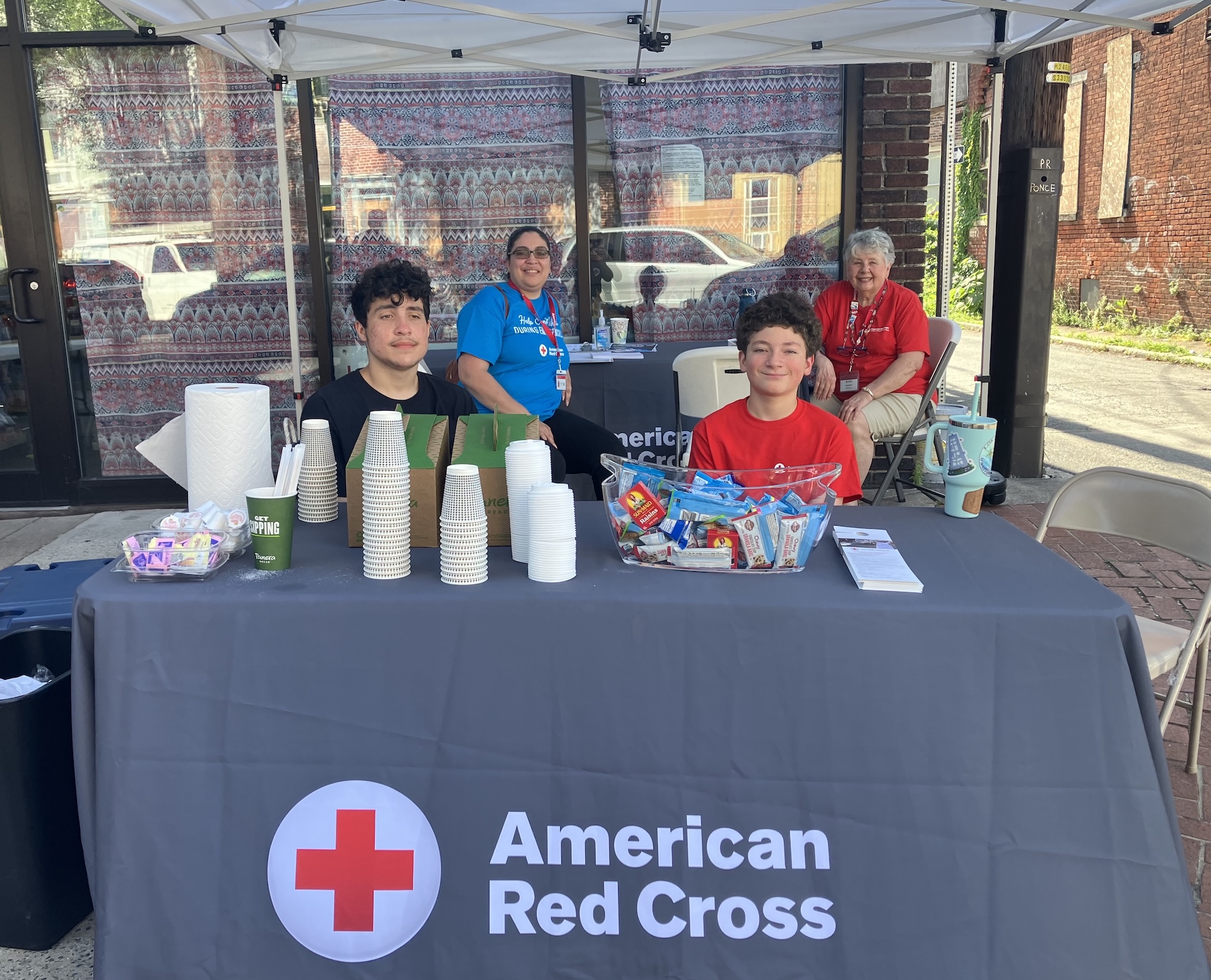 Red Cross volunteers sit at a table under a canopy at a community event. Rosalie Gonzalez, wearing a blue shirt, is seated in the back with another volunteer in red. In the front, her son Landon, in a red shirt, and another young volunteer sit behind a table covered with snacks, cups, and Red Cross materials.