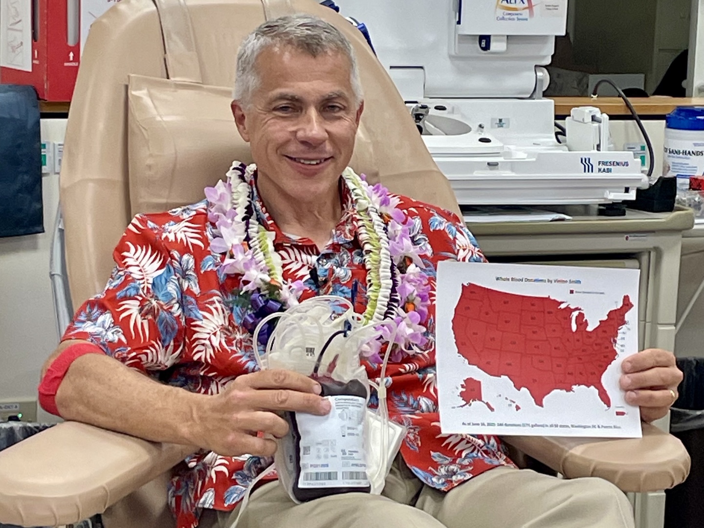 Vinton Smith, wearing a floral shirt and lei, smiles after donating blood in Hawaii. He is holding his blood bag and a map of the United States with all states colored red.