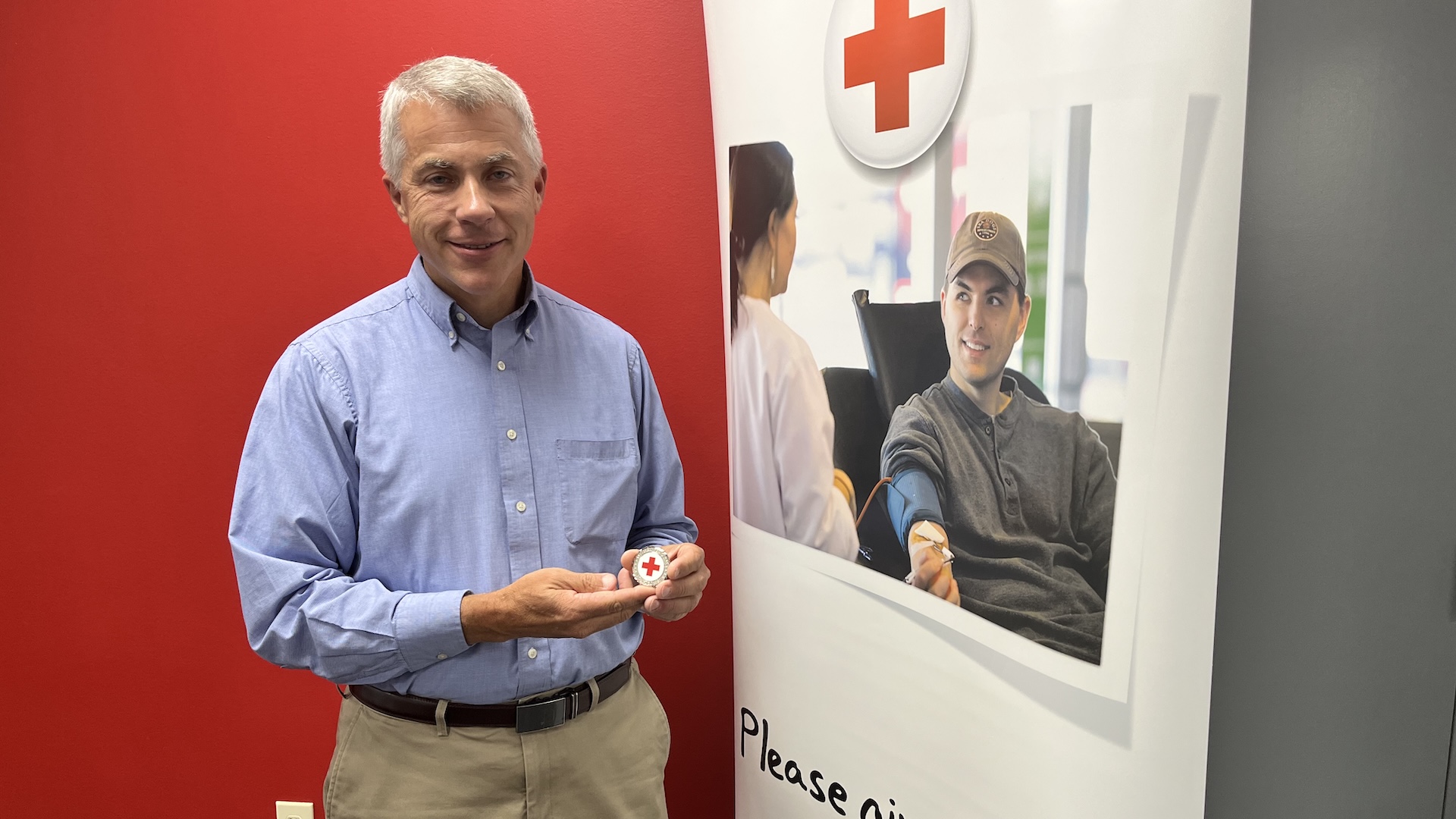 Vinton Smith holds Red Cross Challenge Coin in front of banner showing man giving blood with text "Please give blood".