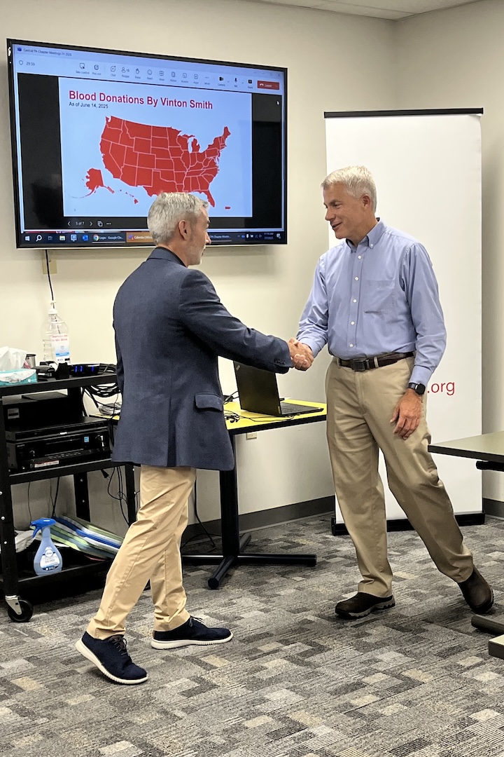 Vinton Smith, on right, shakes hands with Jorge Martinez,  while receiving the American Red Cross of Greater Pennsylvania Challenge Coin. A monitor behind them displays a map of the United States, with each state colored red to represent Vinton's blood donations in each state.