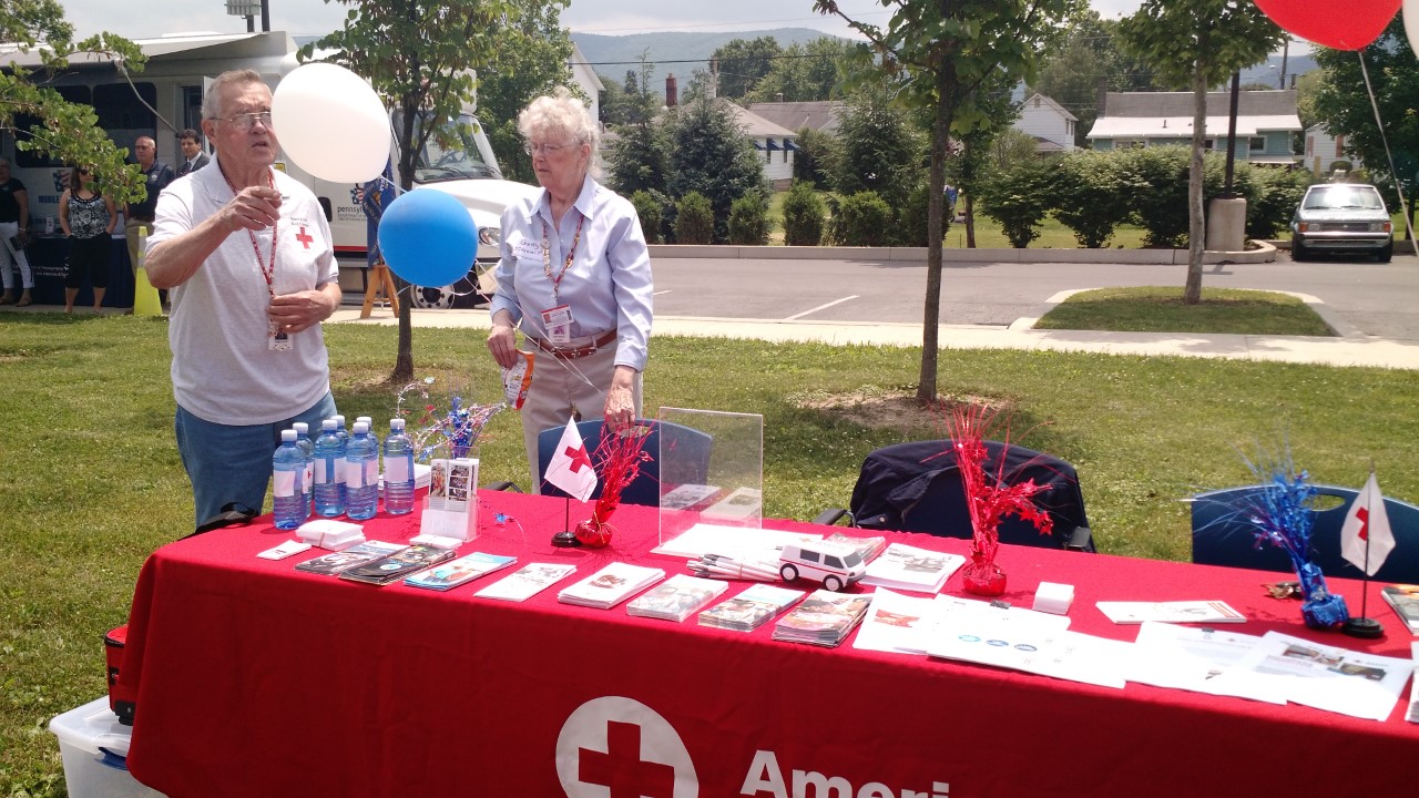 Getty Stewart and another Red Cross volunteer stand behind a table with an American Red Cross tablecloth and various handout during a community event.