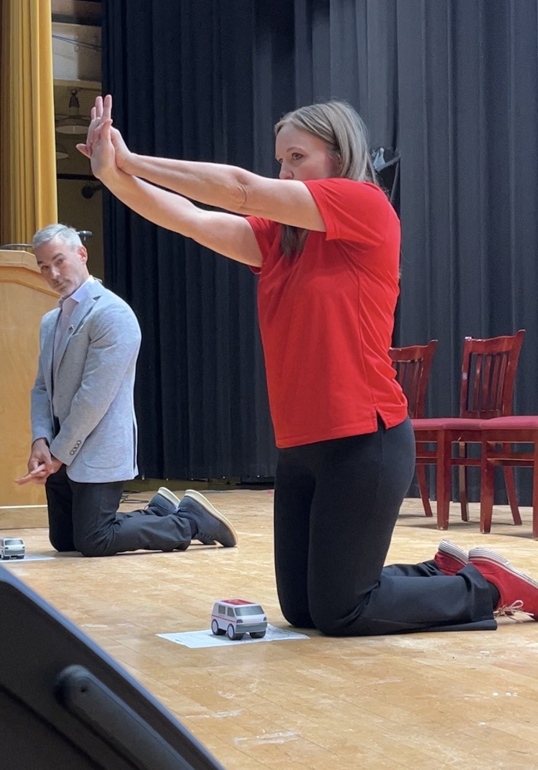 Melinda Rosario kneels and outstretches her arms in front of her, with interlocked fingers, demonstrating the hand positioning for Hands-Only CPR. Jorge Martinez watches from the background.