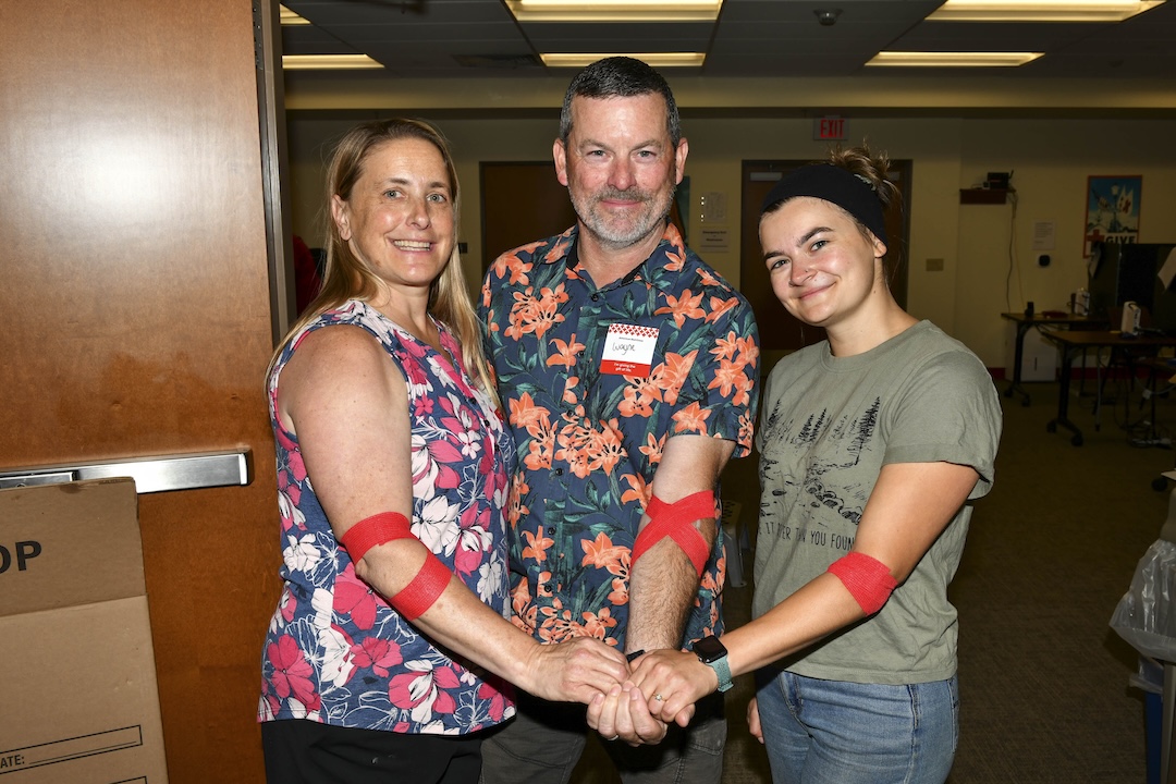 Three adults with arms extended, with a red arm wrap after donating blood