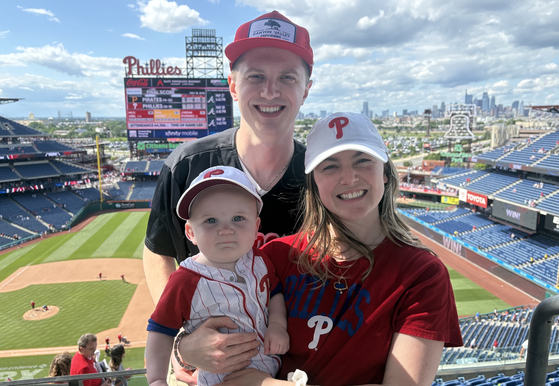 Kylie McCullough, smiles while holding her son Tedddy, with her husband Peter. They are wearing Philadelphia Phillies attire, while the ballpark and city skyline are in the background.