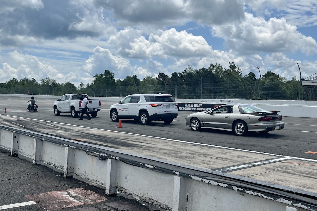 Vehicles, including a motorcycle and a Red Cross SUV, lined up on pit road at Pocono Raceway.