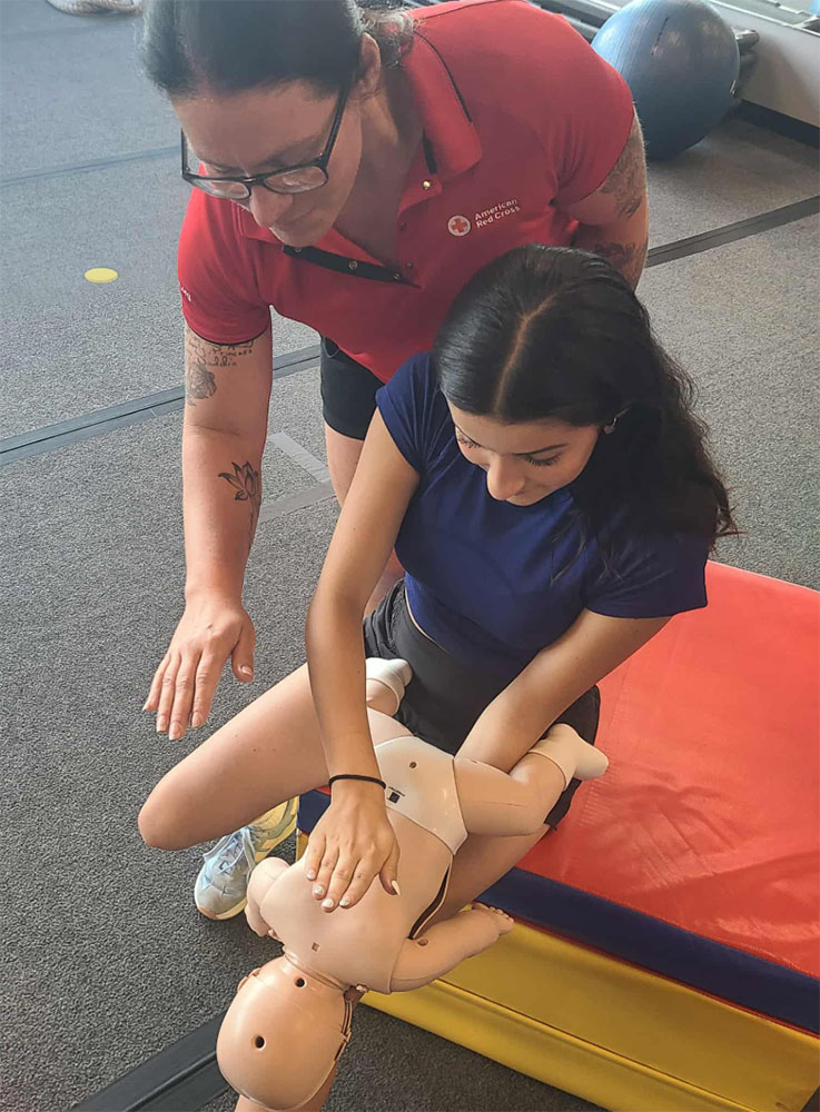 Amber Marchetta standing next to a girl who is holding a CPR manikin baby and practicing on it.