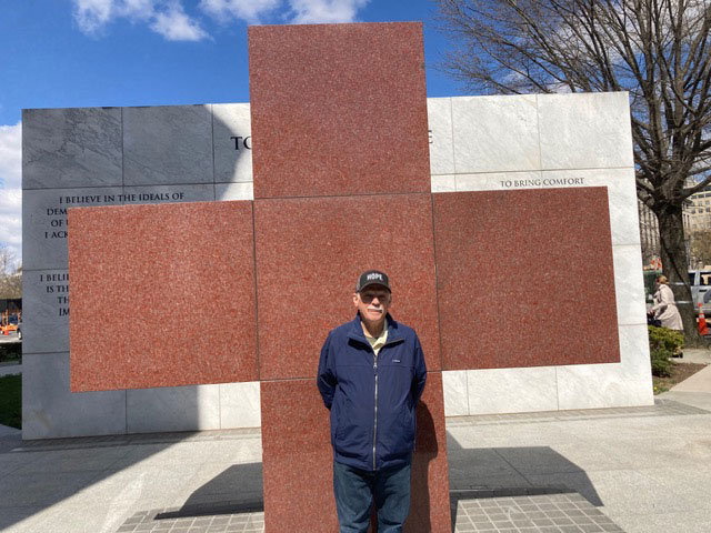 Rich Dansizen, wearing a hat and jacket, stands in front of a large marble Red Cross monument.