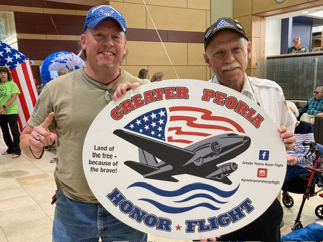 Rich Dansizen and another person holding a large sign that says Greater Peoria Honor Flight with a plane and American flag drawn on it.