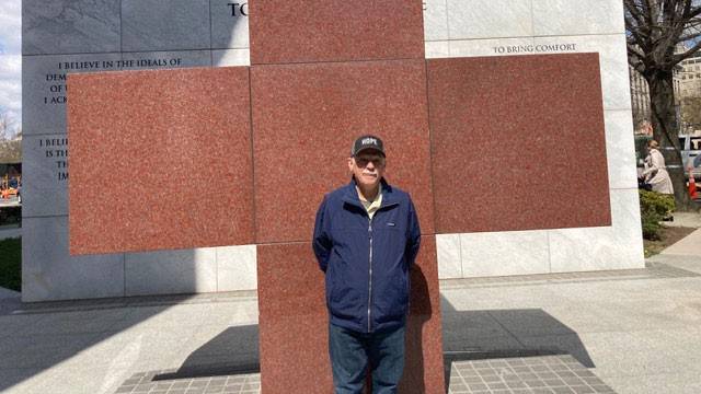 Rich Dansizen, wearing a hat and jacket, stands in front of a large marble Red Cross monument.