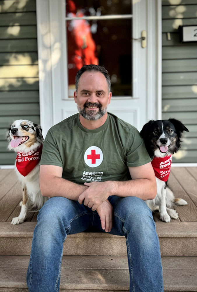 Logan Hamilton wearing a Red Cross shirt and sitting on a porch with service dogs nova and lucy.