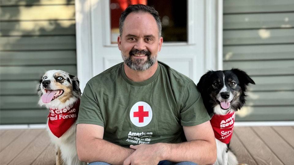 Logan Hamilton wearing a Red Cross shirt and sitting on a porch with service dogs nova and lucy.