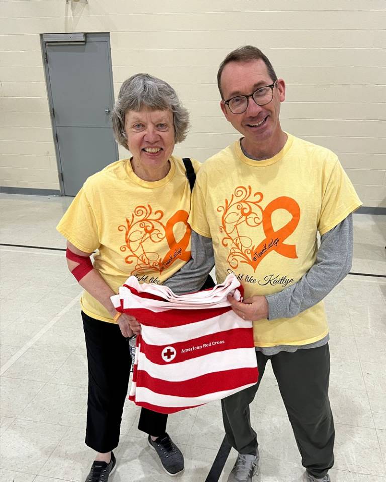 Two people in yellow shirts that read "Fight Like Kaitlyn" smile at the camera. They are holding a red and white beach towel with the American Red Cross logo.