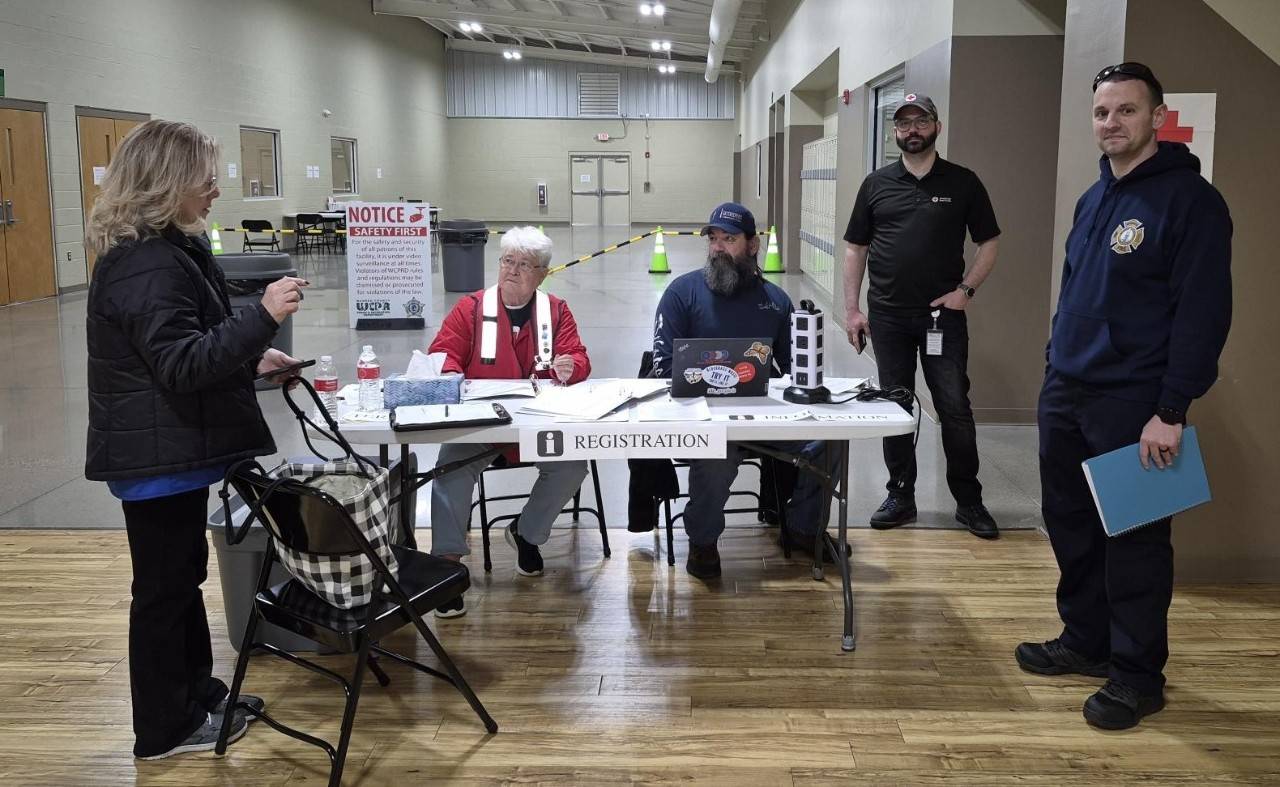 A group of people talk around a white folding table labeled "Registration". One of the people pictured is wearing a Red Cross vest and t-shirt. In the background, there is an open area similar to a gym with a sign that says "Notice, Safety First".