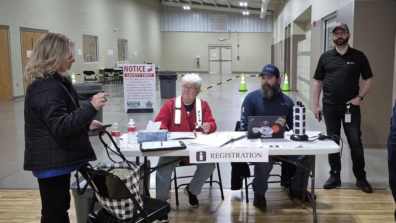 A group of people talk around a white folding table labeled "Registration". One of the people pictured is wearing a Red Cross vest and t-shirt. In the background, there is an open area similar to a gym with a sign that says "Notice, Safety First".