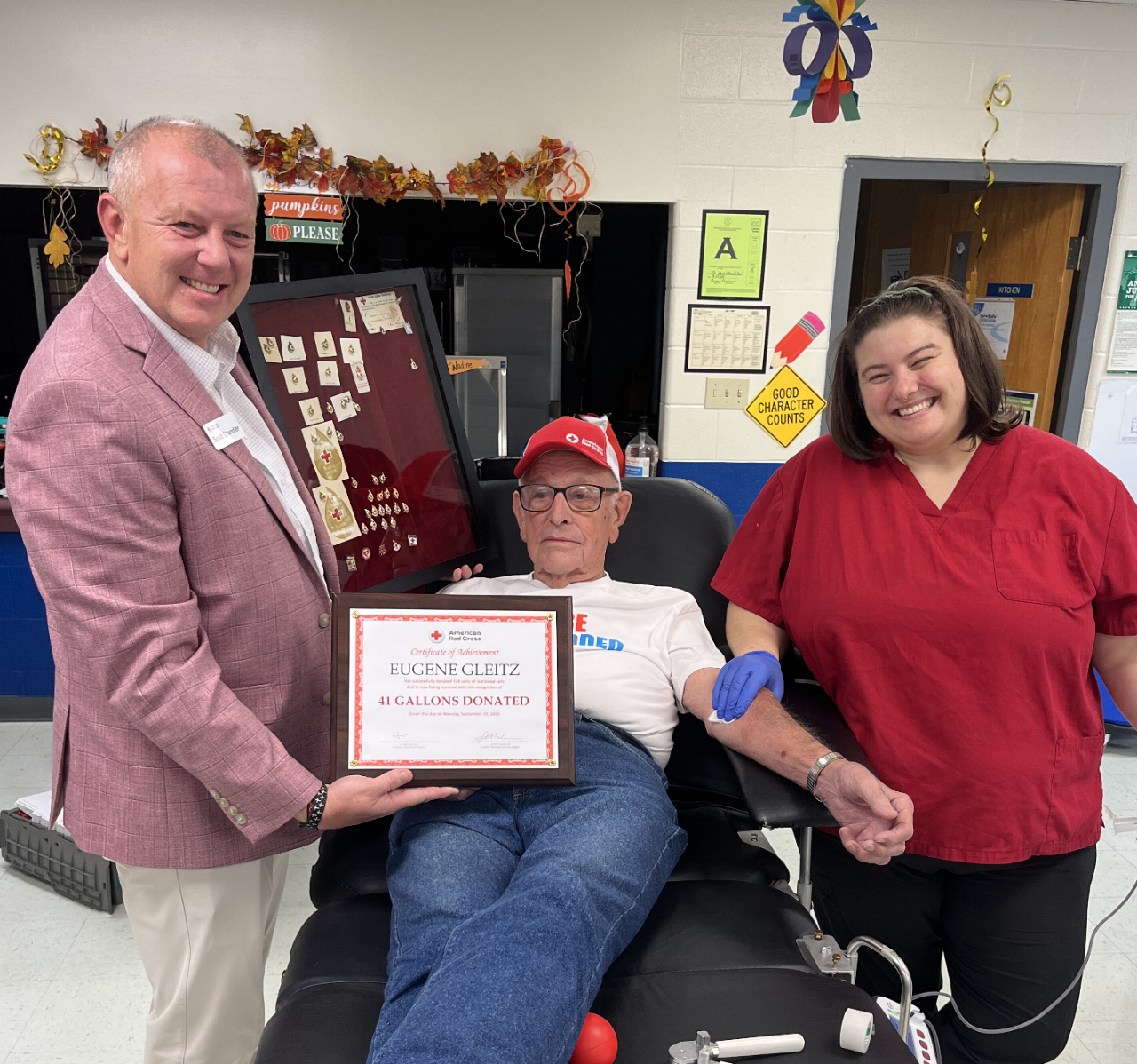 Three people at a Red Cross blood drive. The one in the middle, Eugene Gleitz, is sitting on a blood donation bed after making his 41st gallon donation. The person on the left is holding a pin board of pins Gleitz has earned over the course of his life and a plaque that readds "41 Gallons Donated".