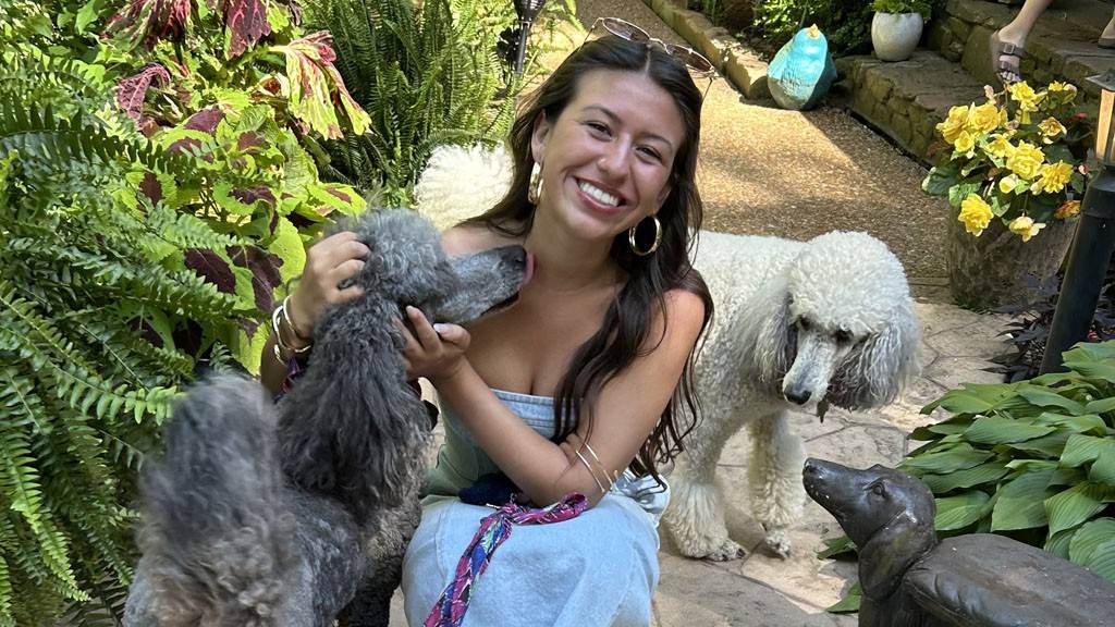 A young woman crouches next to a poodle in an outdoor setting. She is smiling at the camera.