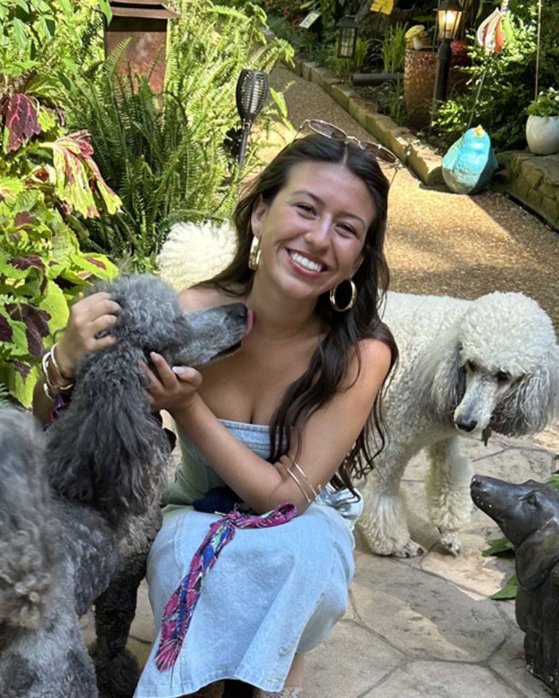 A young woman crouches with a poodle in an outdoor setting. She is smiling at the camera.