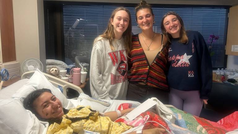 Four young people smile at the camera. The one on the left is lying in a hospital bed. The one on the middle left is wearing a Western Kentucky University sweatshirt, and the one on the far right is wearing a shirt that says Kappa Delta.