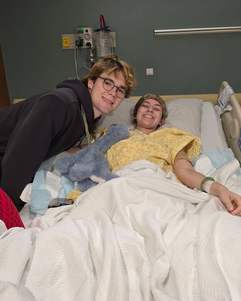 Two young people smile at the camera. The one on the right is laying in a hospital bed in a yellow hospital gown.