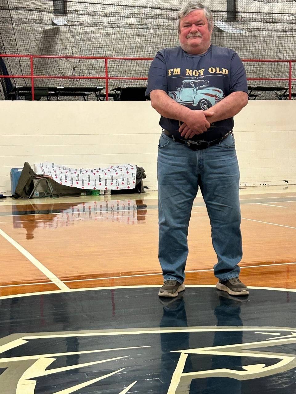 A man in a shirt that reads "I'm not old" stands in a school gym with a Red Cross cot and blanket in the background. There is an image of the Monroe County High School Mascot, a falcon, on the ground in front of him.