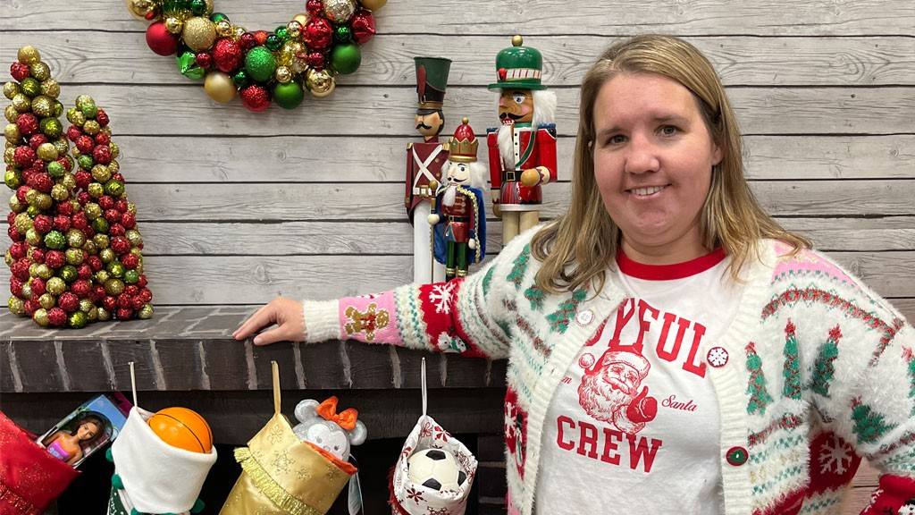 A Red Cross volunteer in a festive holiday sweater smiles. They are standing in front of a faux fireplace with holiday decorations, stockings and a Red Cross logo on the hearth.