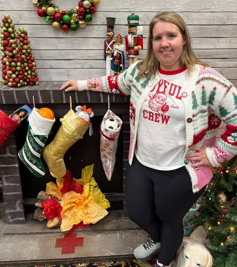 A Red Cross volunteer in a festive holiday sweater smiles. They are standing in front of a faux fireplace with holiday decorations, stockings and a Red Cross logo on the hearth.